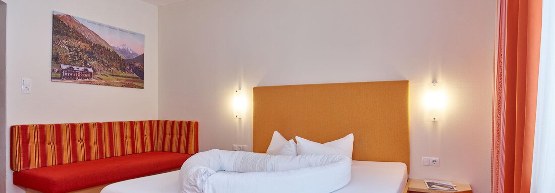 Ein gemütliches Schlafzimmer mit einem Doppelbett und einer roten Sitzbank. Die Wände sind hell, und es gibt ein Bild eines Bergs im Hintergrund. - Hotel Vent - Vent