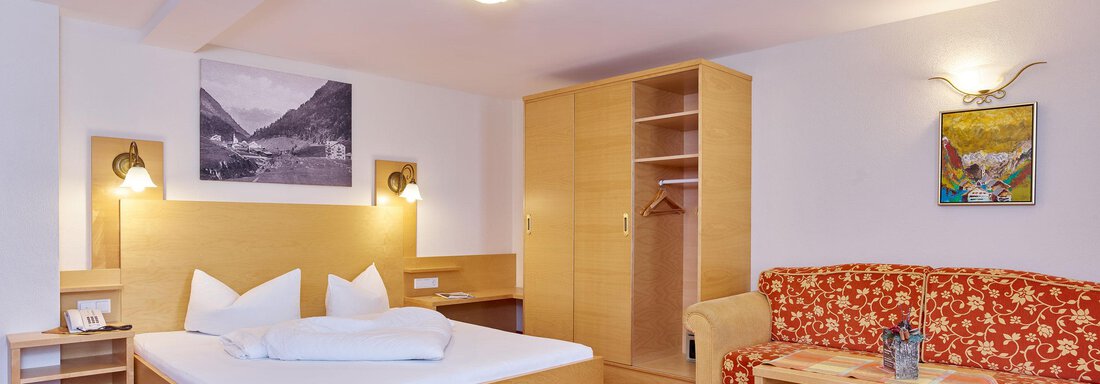 Ein modernes Hotelzimmer mit einem Doppelbett, einer Sitzgruppe und einem Kleiderschrank. Die Wände sind hell und es gibt dekorative Bilder. - Hotel Vent - Vent