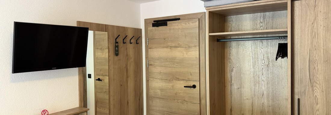 A modern hotel room with a television on the wall. The wardrobe is made of wood and provides space for clothes and personal belongings. - Mein Weisskugel - Vent