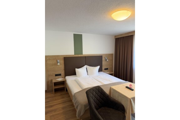 A modern hotel room with a large bed and a wood panel. Next to the bed, there is a small table and a chair. - Mein Weisskugel - Vent