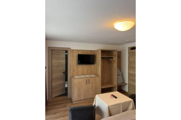 A modern room with wooden furniture and a television on the wall. There is a table and a wardrobe as well as a door to a bathroom. - Mein Weisskugel - Vent