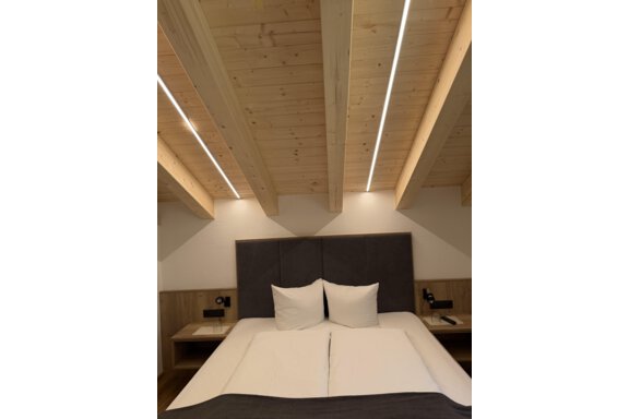 A modern bedroom with a large bed and two pillows. The wooden ceiling has visible beams and soft lighting. - Mein Weisskugel - Vent