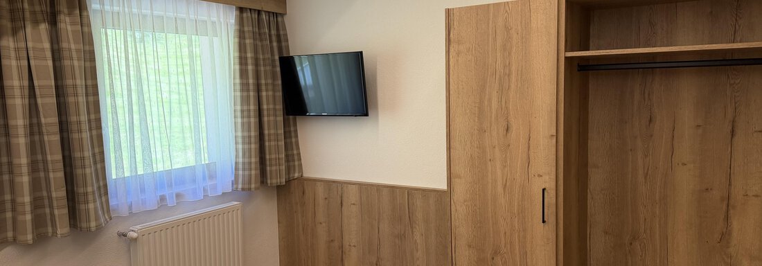 A modern bedroom with wood paneling and a television on the wall. The room has a window with curtains and an open wardrobe. - Mein Weisskugel - Vent