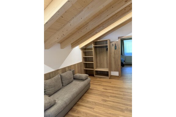 A cozy room with a gray sofa and a wooden ceiling. There is a shelf and a window that lets natural light in. - Mein Weisskugel - Vent