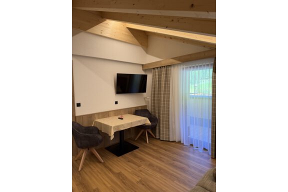 A cozy living area with a table, two chairs, and a television on the wall. Large windows let in plenty of light and offer a view outside. - Mein Weisskugel - Vent