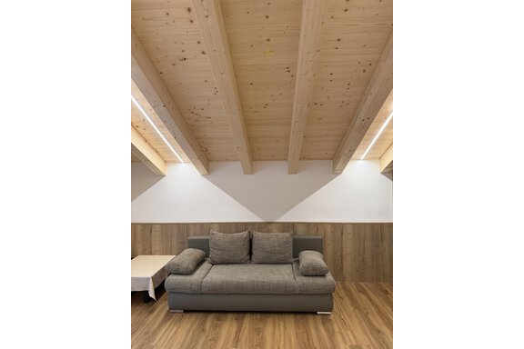 A modern living room with a gray sofa and a light wooden floor. The ceiling is clad with wooden panels and has large windows that let in natural light. - Mein Weisskugel - Vent