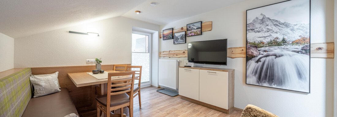 Wohnraum Bergzeit Five | © Brunner Johannes, Michaela Jörg (CC C) A cozy living space with modern furnishings. There is a dining table, a sofa, and a television, complemented by a mountain picture on the wall. - Garni Ötztal ROOMS & APARTS - Zwieselstein