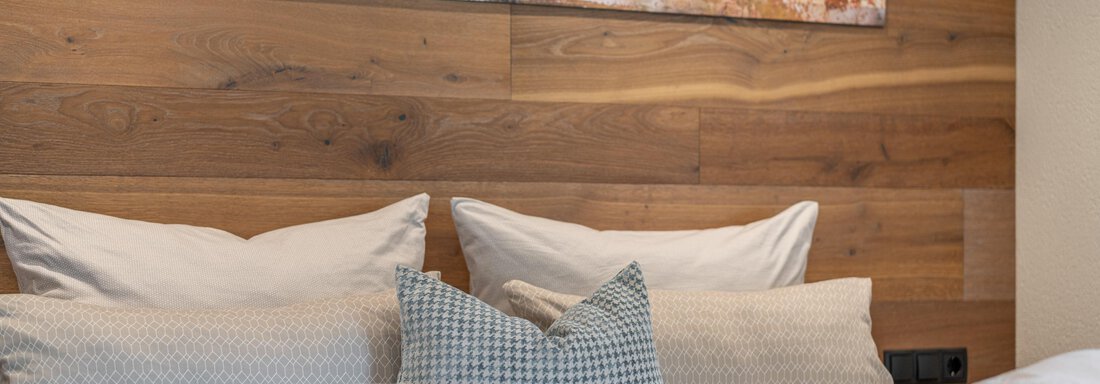 A cozy bedroom with a wooden panel and several cushions on the bed. A modernly designed picture hangs on the wall. - Garni Ötztal ROOMS & APARTS - Zwieselstein