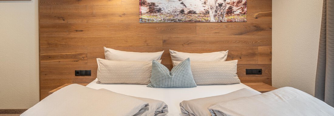 A modern bedroom with a double bed, cozy cushions, and a wooden panel wall. A beautiful picture of mountains hangs on the wall. - Garni Ötztal ROOMS & APARTS - Zwieselstein
