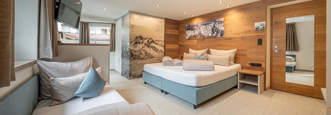 Bergzeit Trio | © Brunner Johannes, Michaela Jörg (CC C) A modern room with a large bed and a seating area. The walls are adorned with wooden paneling and there is a window with a view outside. - Garni Ötztal ROOMS & APARTS - Zwieselstein