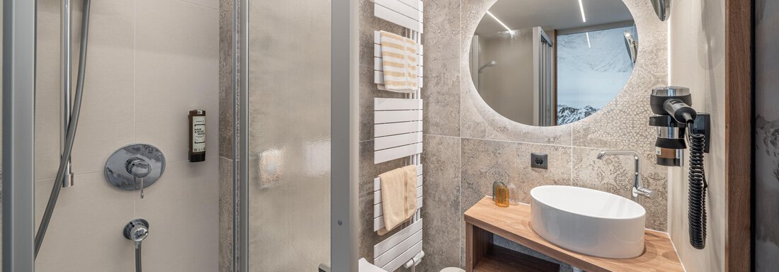 A modern bathroom with a shower, a round mirror, and an elegant sink. The walls are in neutral tones and there are towels nearby. - Garni Ötztal ROOMS & APARTS - Zwieselstein