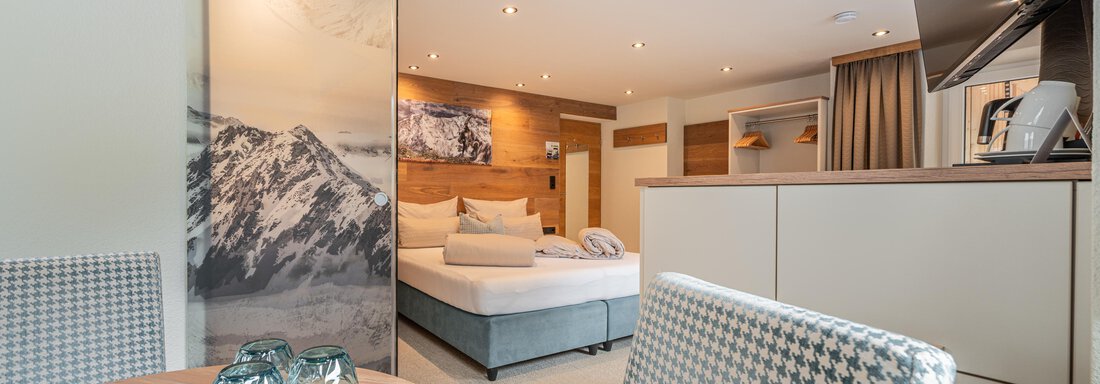 A modern room with a cozy bed and a table. The walls are light and there is a large window pane with a view of mountains. - Garni Ötztal ROOMS & APARTS - Zwieselstein