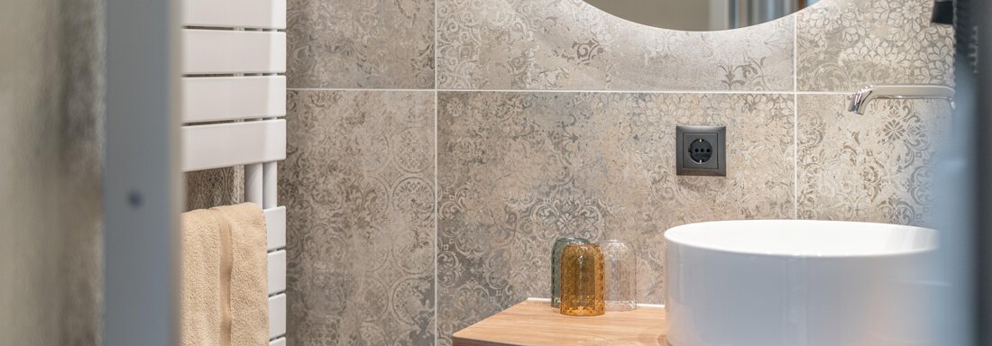 A modern bathroom with a round mirror and an elegant sink. The walls are decorated with stylish tiles and there are towels nearby. - Garni Ötztal ROOMS & APARTS - Zwieselstein
