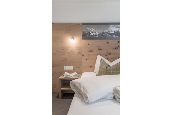 Ein gemütliches Schlafzimmer mit einem Holzschrank und einem modernen Nachttisch. Auf dem Bett liegen Kissen in sanften Farben und ein aufgeschlagenes Buch. - Garni Ötztal ROOMS & APARTS - Zwieselstein