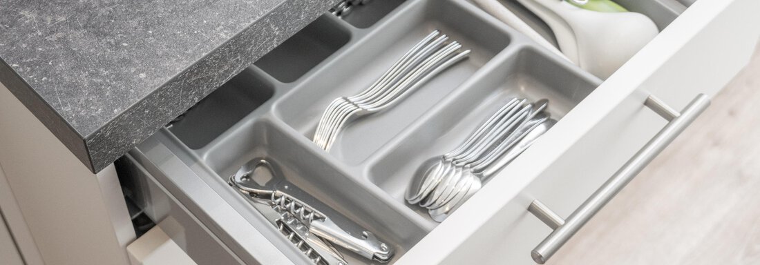 An open drawer in the kitchen displays neatly arranged cutlery and kitchen utensils. The drawer bottom is gray and the countertop is black. - Garni Ötztal ROOMS & APARTS - Zwieselstein
