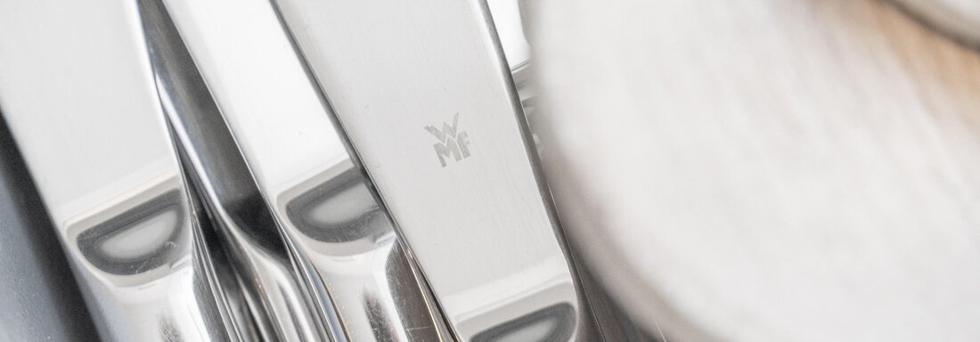 A set of shiny cutlery lies neatly next to each other. In the foreground, a wooden spoon is visible. - Garni Ötztal ROOMS & APARTS - Zwieselstein
