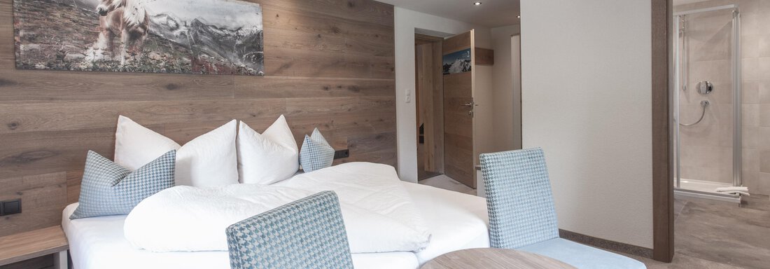 A modern hotel room with a large bed and comfortable chairs. The walls are made of wood and there is an adjoining bathroom. - Garni Ötztal ROOMS & APARTS - Zwieselstein