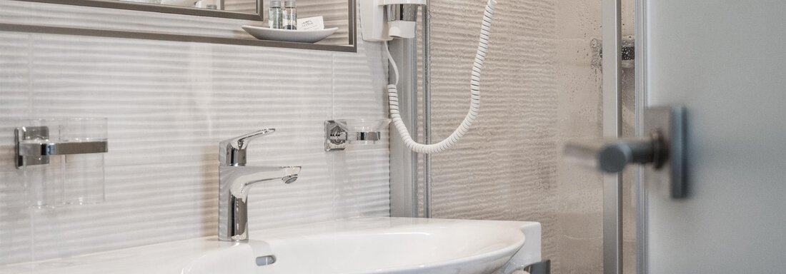 A modern bathroom with a sink, a handheld shower, and a large mirror. The walls are brightly tiled, and a towel is ready. - Garni Ötztal ROOMS & APARTS - Zwieselstein