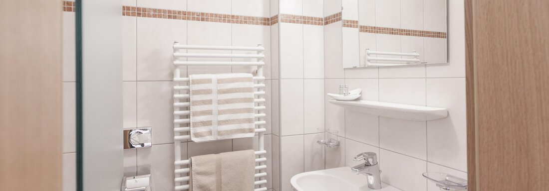 A modern bathroom with white tiles and a mirror. Towels hang on a radiator. - Garni Ötztal ROOMS & APARTS - Zwieselstein