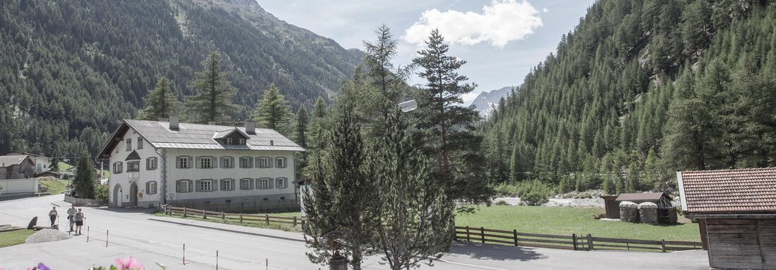 A picturesque mountain landscape with green hills and a clear road. In the foreground stands a traditional building surrounded by trees. - Garni Ötztal ROOMS & APARTS - Zwieselstein
