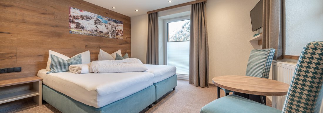 A modern hotel room with a double bed and a small table. Large windows provide plenty of daylight. - Garni Ötztal ROOMS & APARTS - Zwieselstein