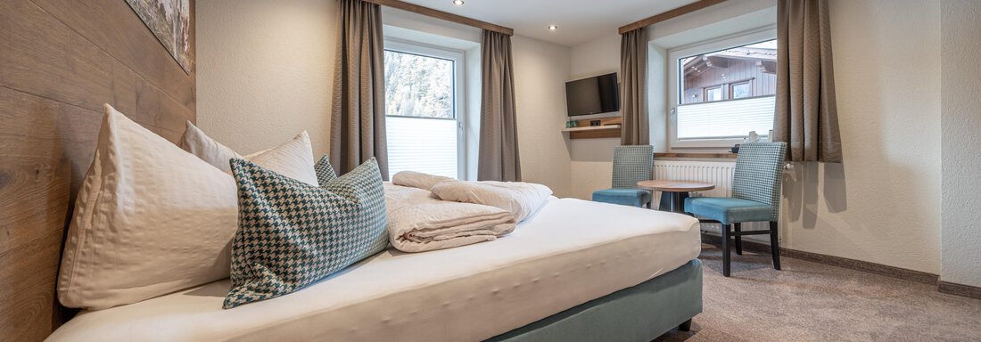 A cozy hotel room with a large bed and modern furniture. There are windows with curtains and a small table with chairs. - Garni Ötztal ROOMS & APARTS - Zwieselstein