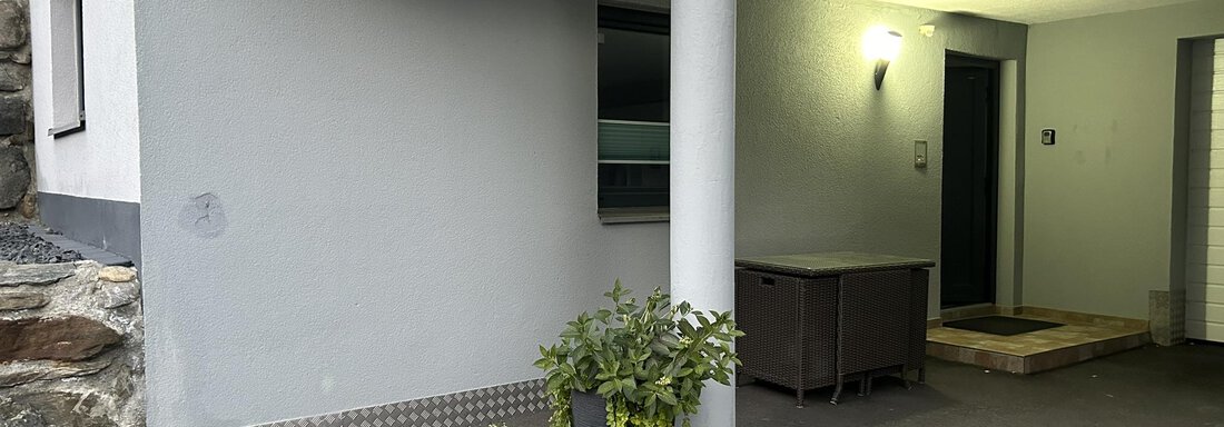 Ein moderner Garagenbereich mit einem Eingang und zwei Töpfen mit Pflanzen. Die Wände sind in einem hellen Grau gehalten und es gibt eine Außenbeleuchtung. - Haus Praxmarer - Zwieselstein