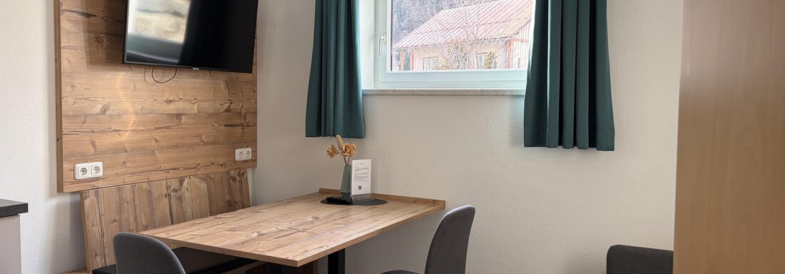 Ein modernes Zimmer mit einem Essbereich. Der Tisch ist aus Holz, und es gibt einen Fernseher an der Wand sowie blaue Vorhänge. - Haus Praxmarer - Zwieselstein